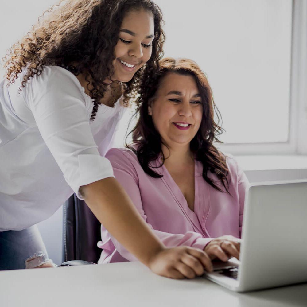 A smiling mother working at a laptop while her happy daughter stands next to her helping