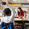 A teacher in a red top addresses a child with their hand up in a classroom