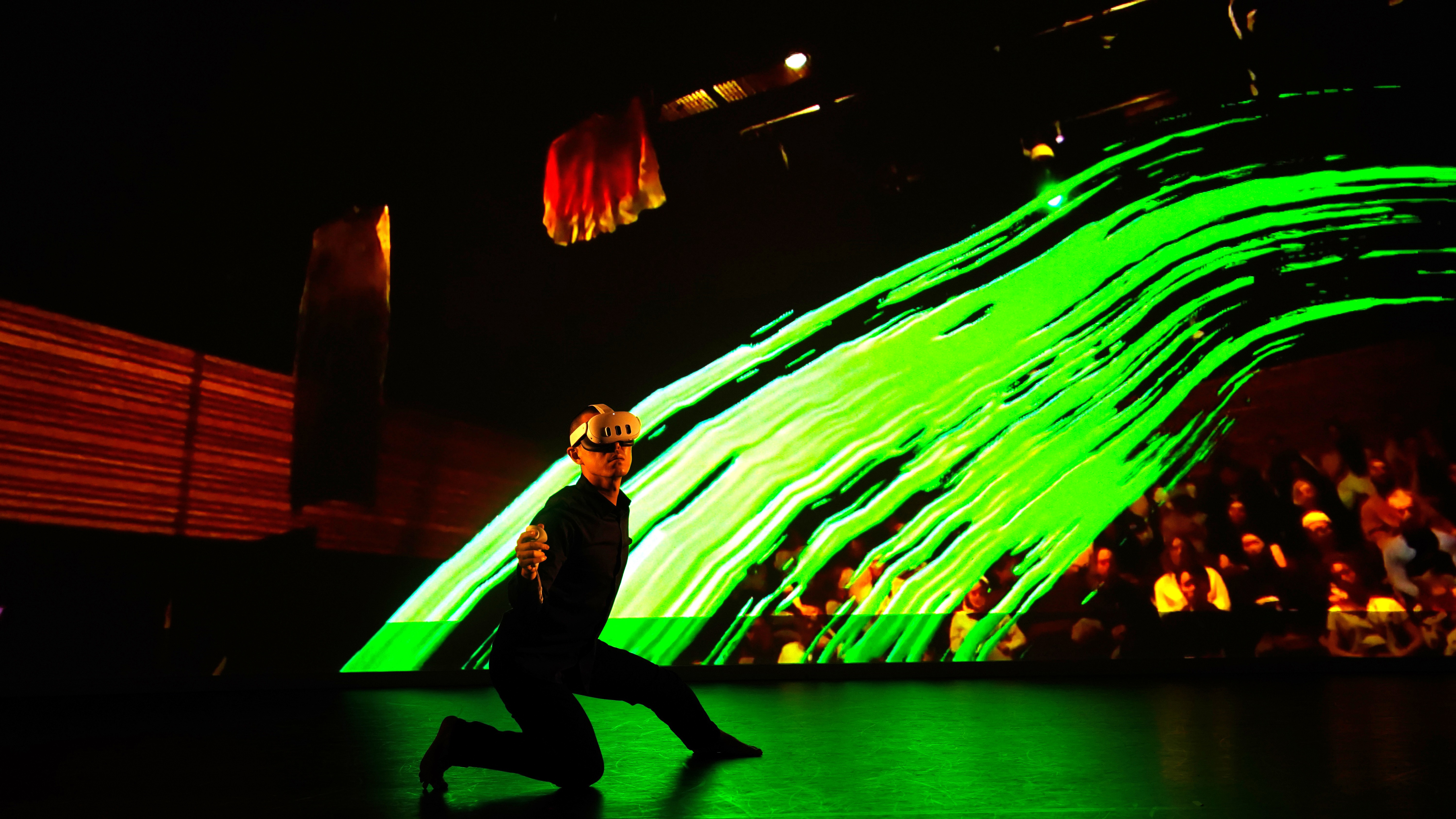 A performer kneels on stage in front of a screen showing the audience and a splash of green colour