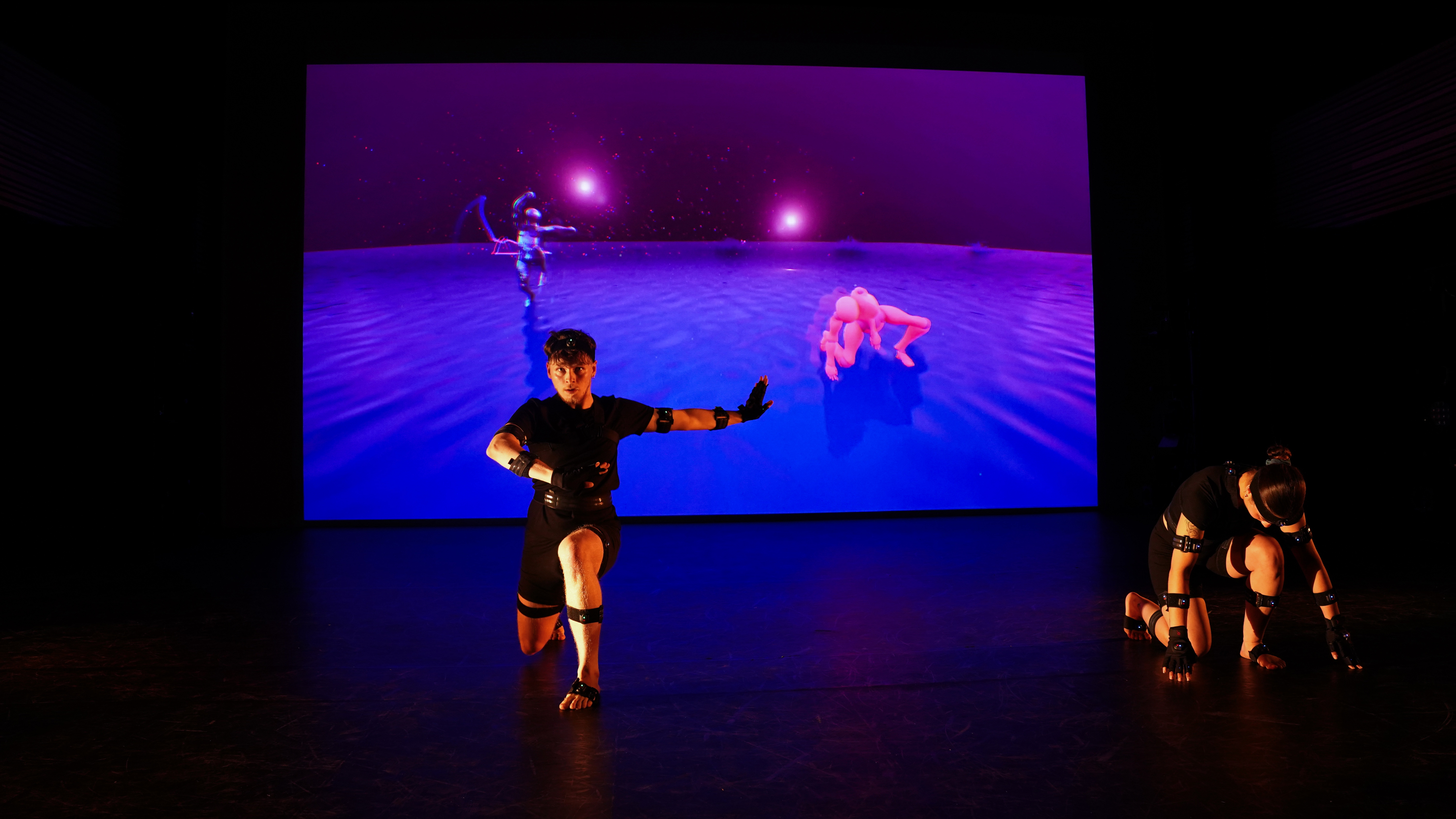 A performer in motion capture equipment poses on a stage in front of a screen showing a digital figure
