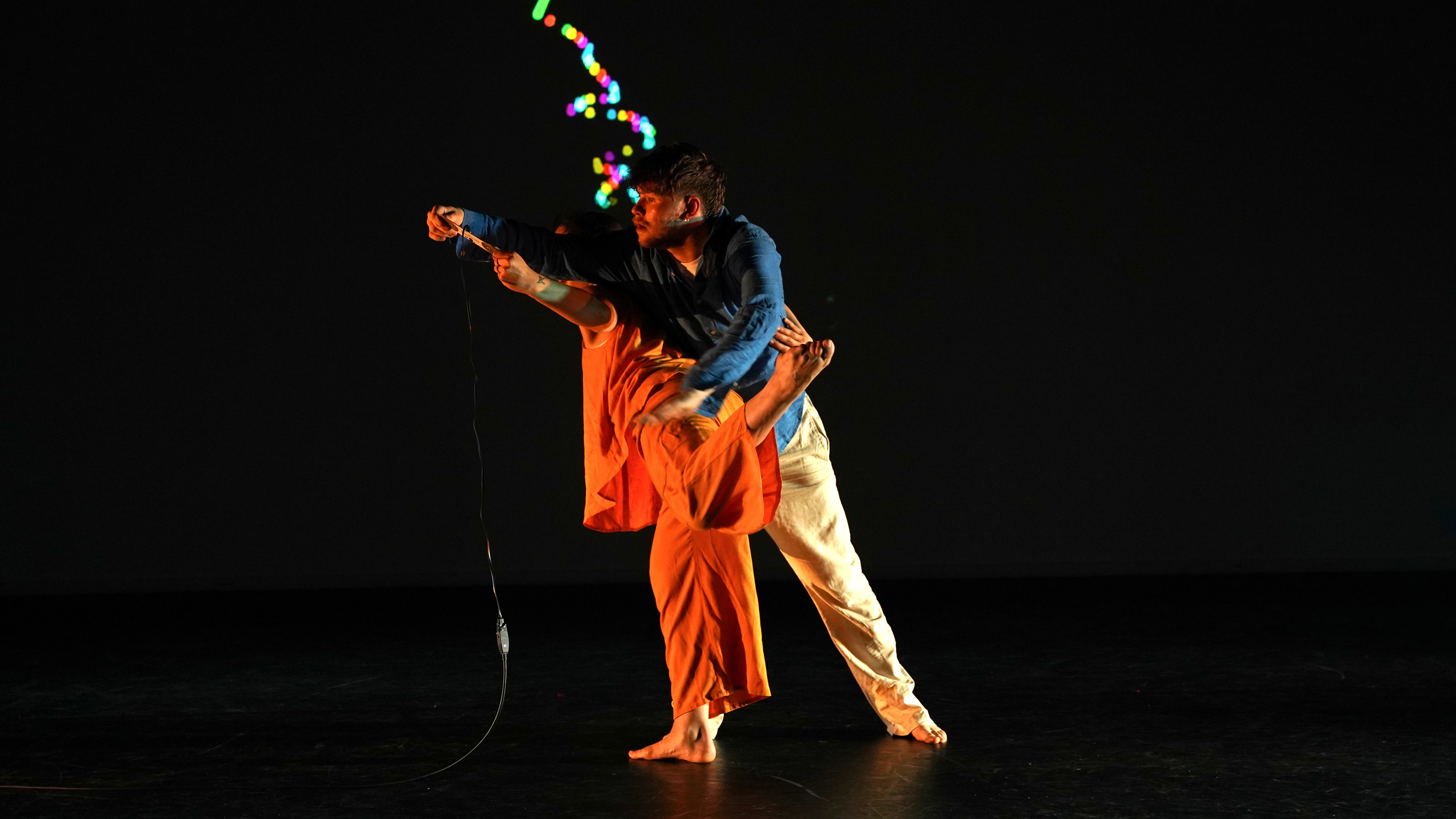 Two performers lean forwards with arms outstretched against a screen showing a spiral of colours