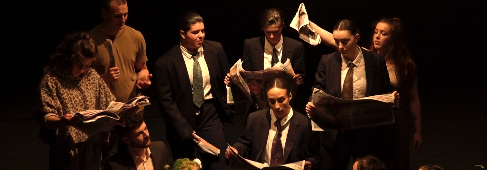 A group of performers in suits read newspapers on a dark stage, dramatically lit from above by a spotlight.