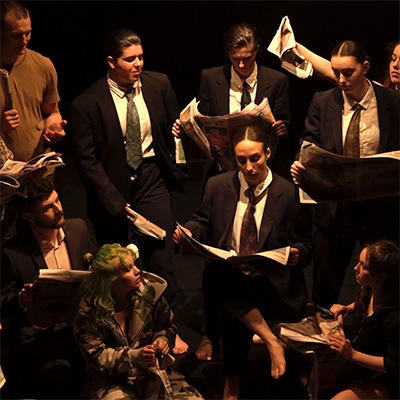 A group of performers in suits read newspapers on a dark stage, dramatically lit from above by a spotlight.