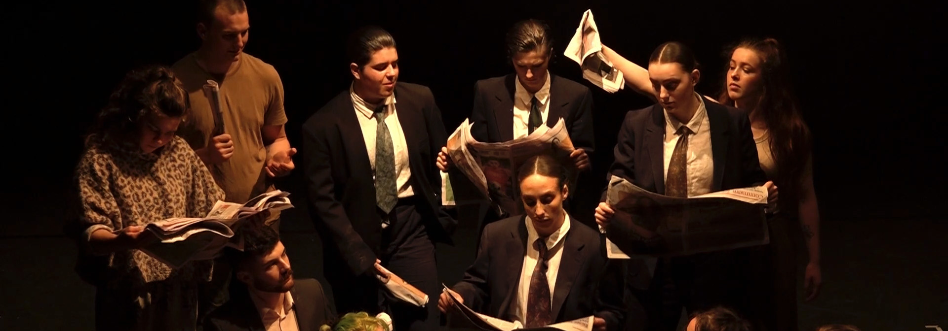 A group of performers in suits read newspapers on a dark stage, dramatically lit from above by a spotlight.