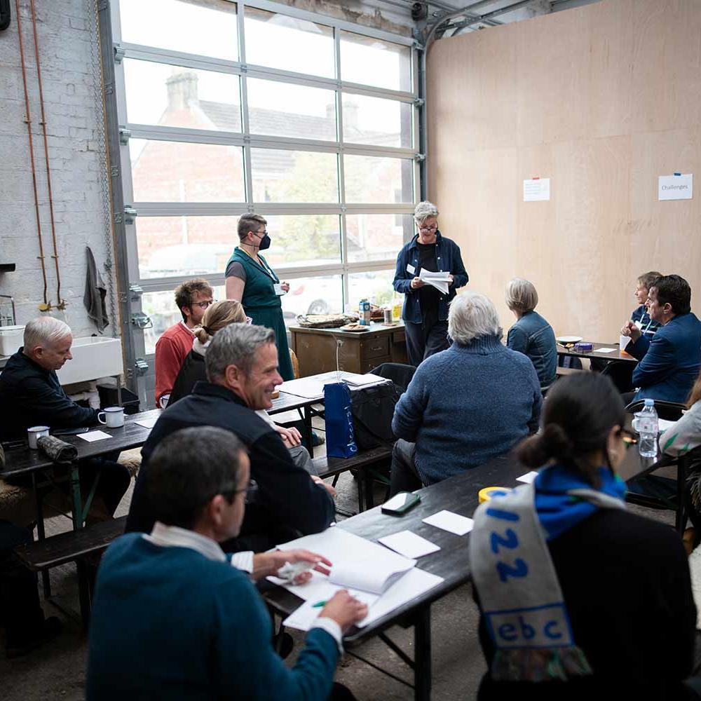 Photograph of room filled with people. Someone is at the front giving a speech