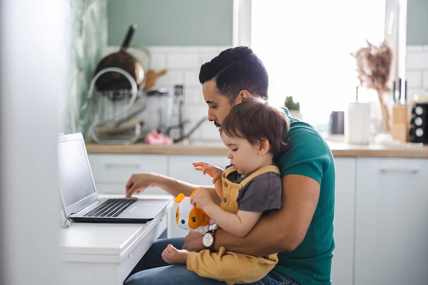 A person holding a toddler sits at a kitchen table and uses a laptop