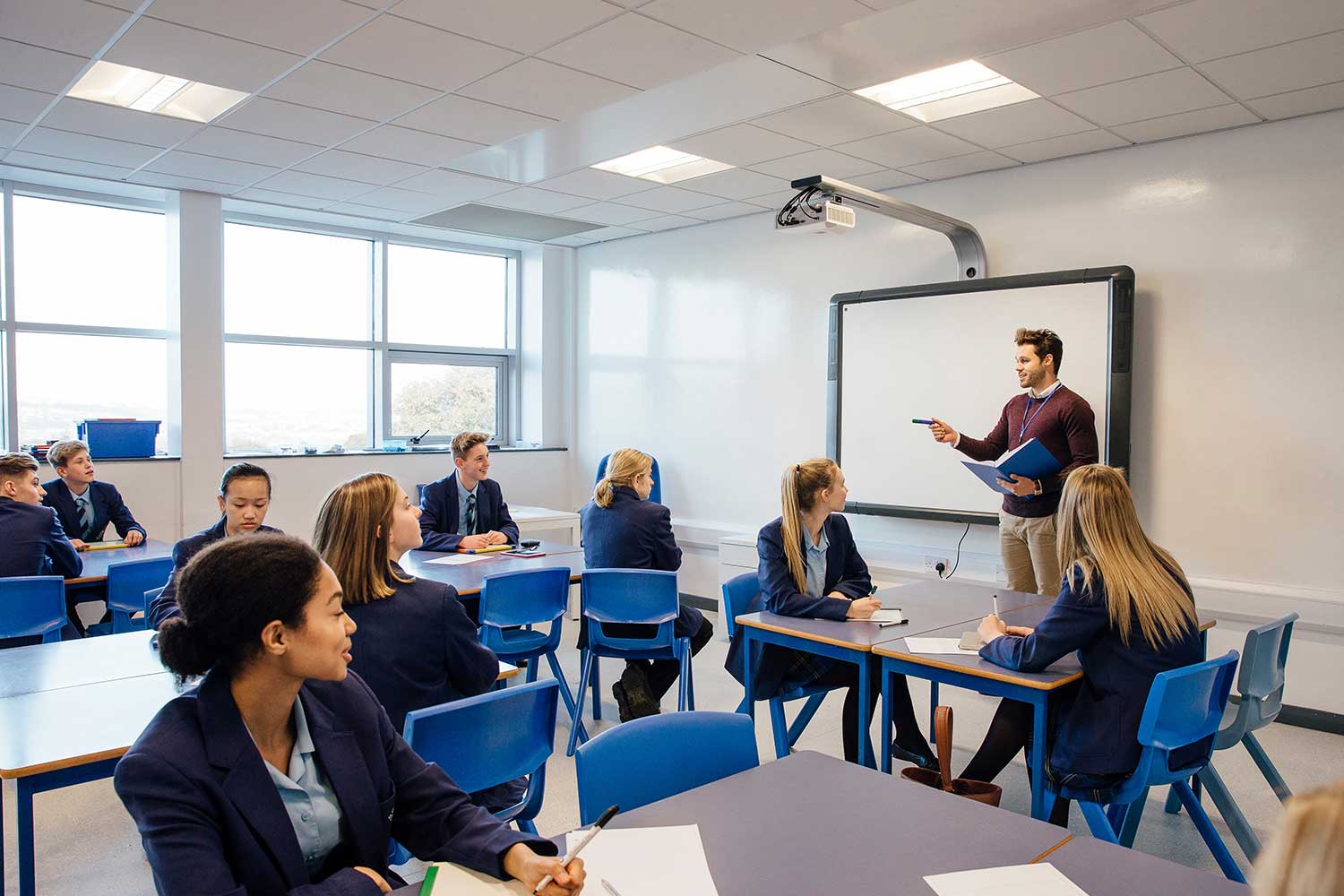 A teacher stands at the front of a class and gestures to one of the pupils to speak