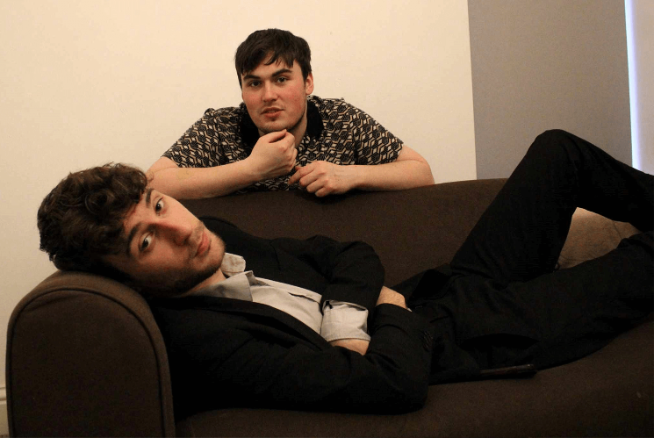 A student lies on a sofa. Behind him an another student leans on the sofa.