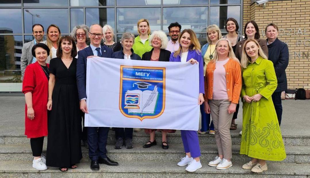 A large group of people stand outside a modern building, holding up a flag with the university's crest on it