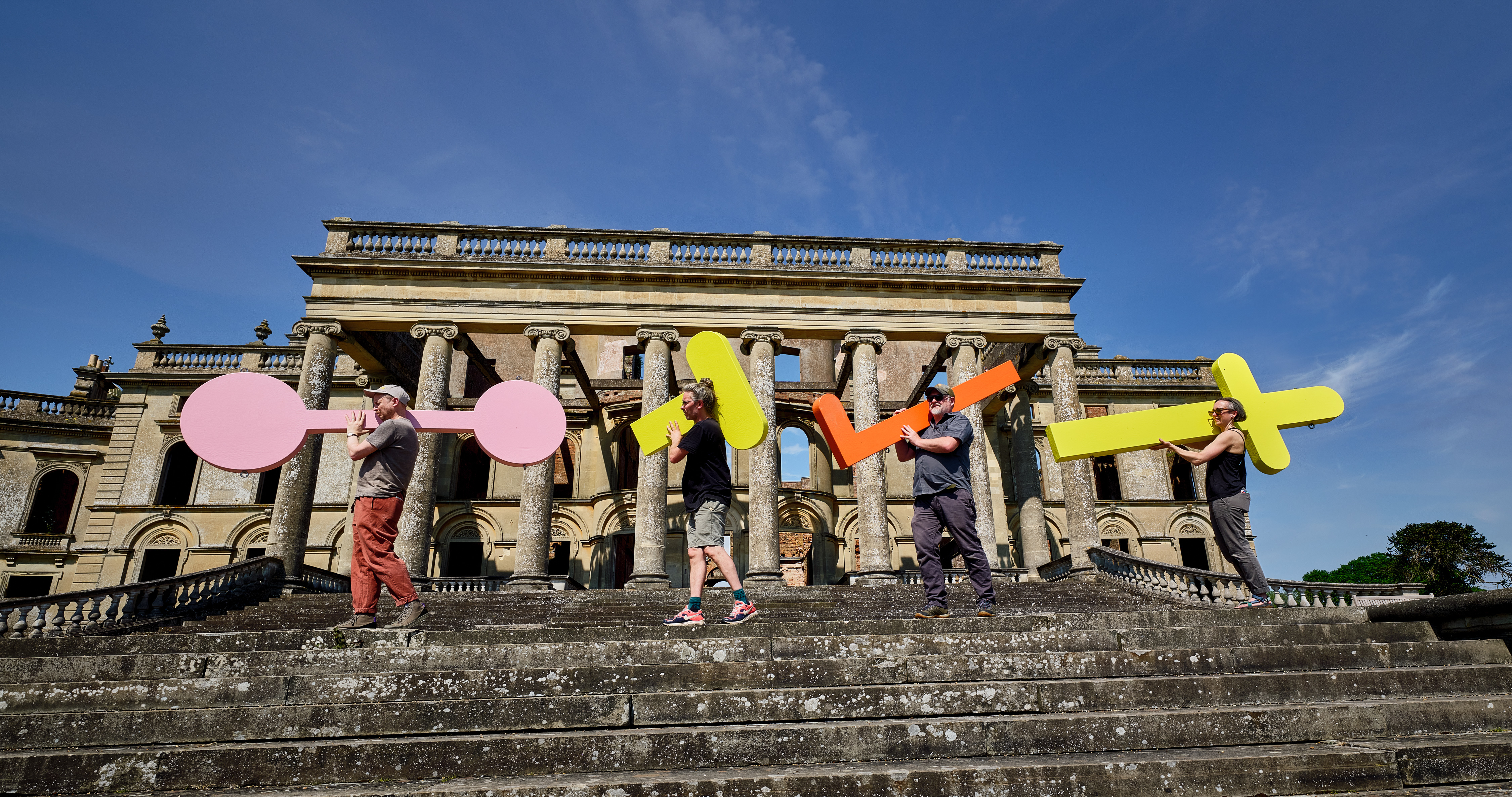 An art installation outside a large grand building with people carrying large colourful shaped letters