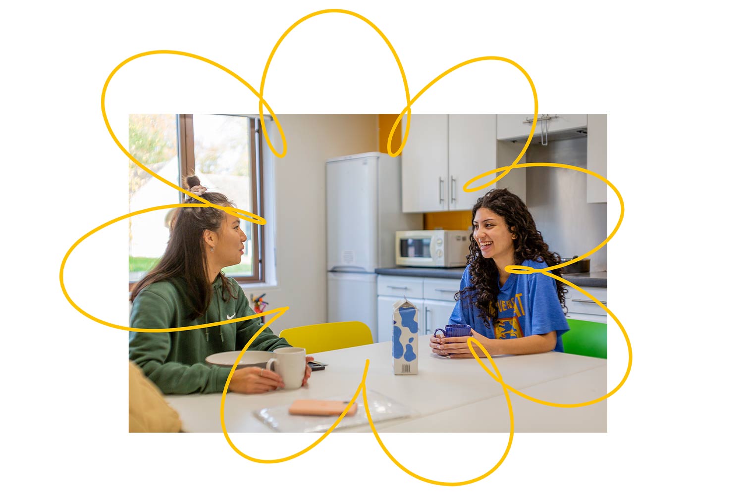 Two students sat at a table in a brightly lit kitchen area with mugs and bowls.