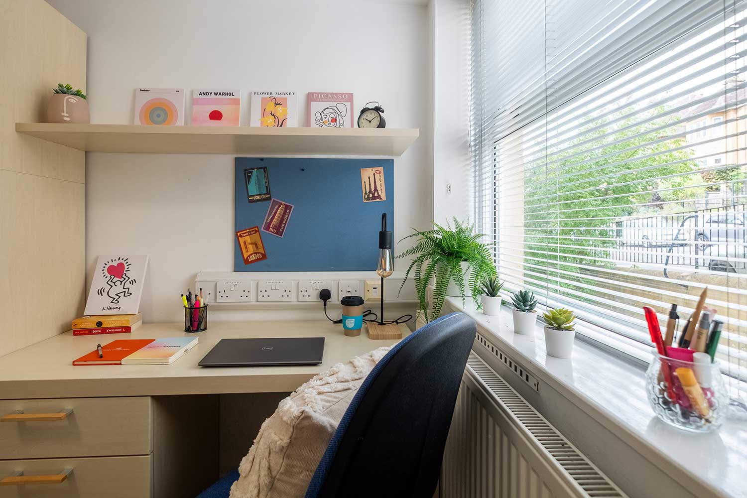 A desk, desk chair and noticeboard next to a window with a blind
