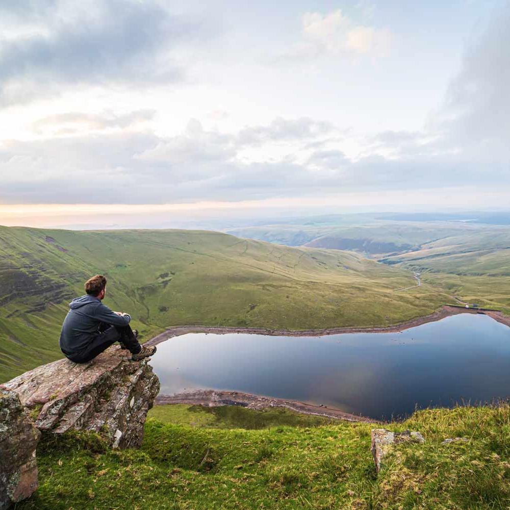 Person in blue hoodie sitting on a rock overlooking lake and mountain view