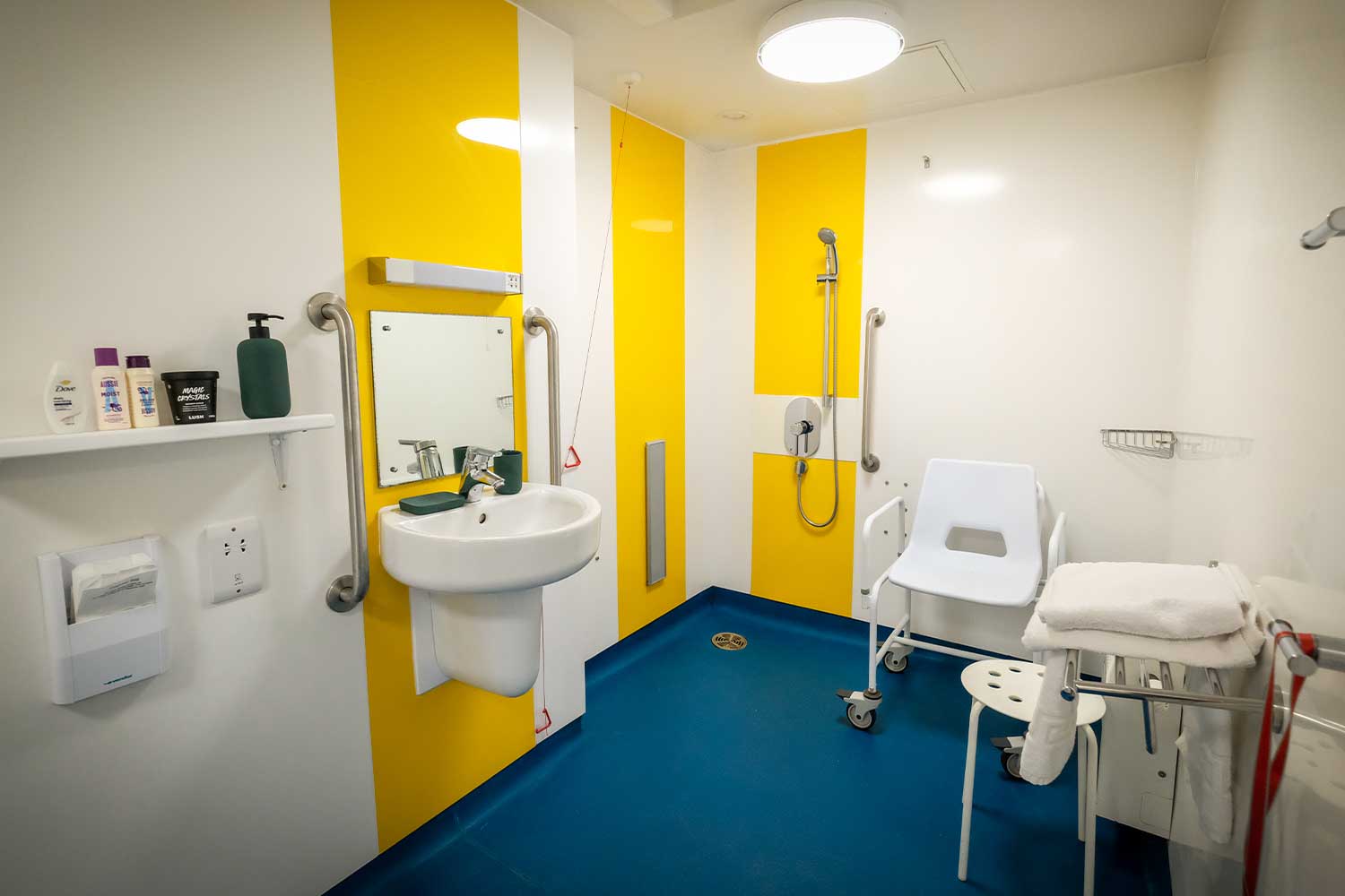 A large bathroom with additional handrails, a walk-in shower on a level surface and a wheeled chair suitable for use in the shower