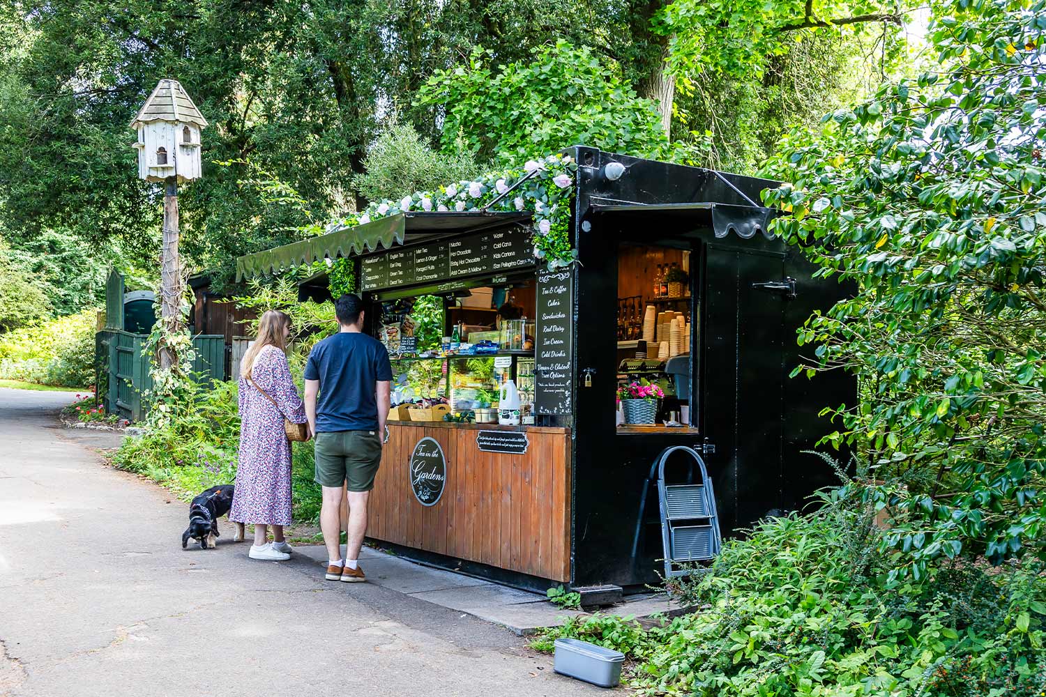 Two people at an outdoor cafe kiosk surrounded by green trees and shrubs