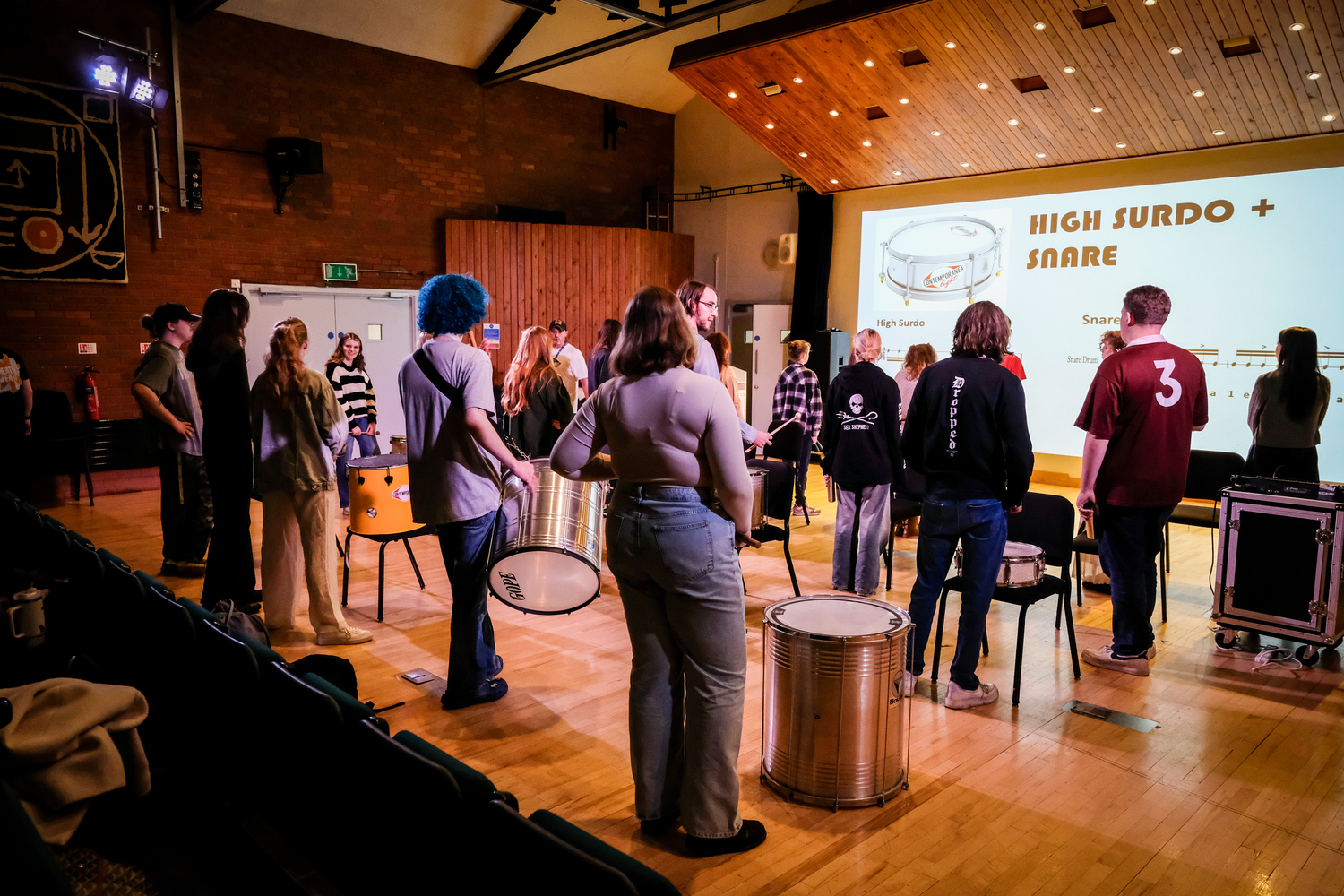 A percussion workshop where people with instruments stand looking at a speaker