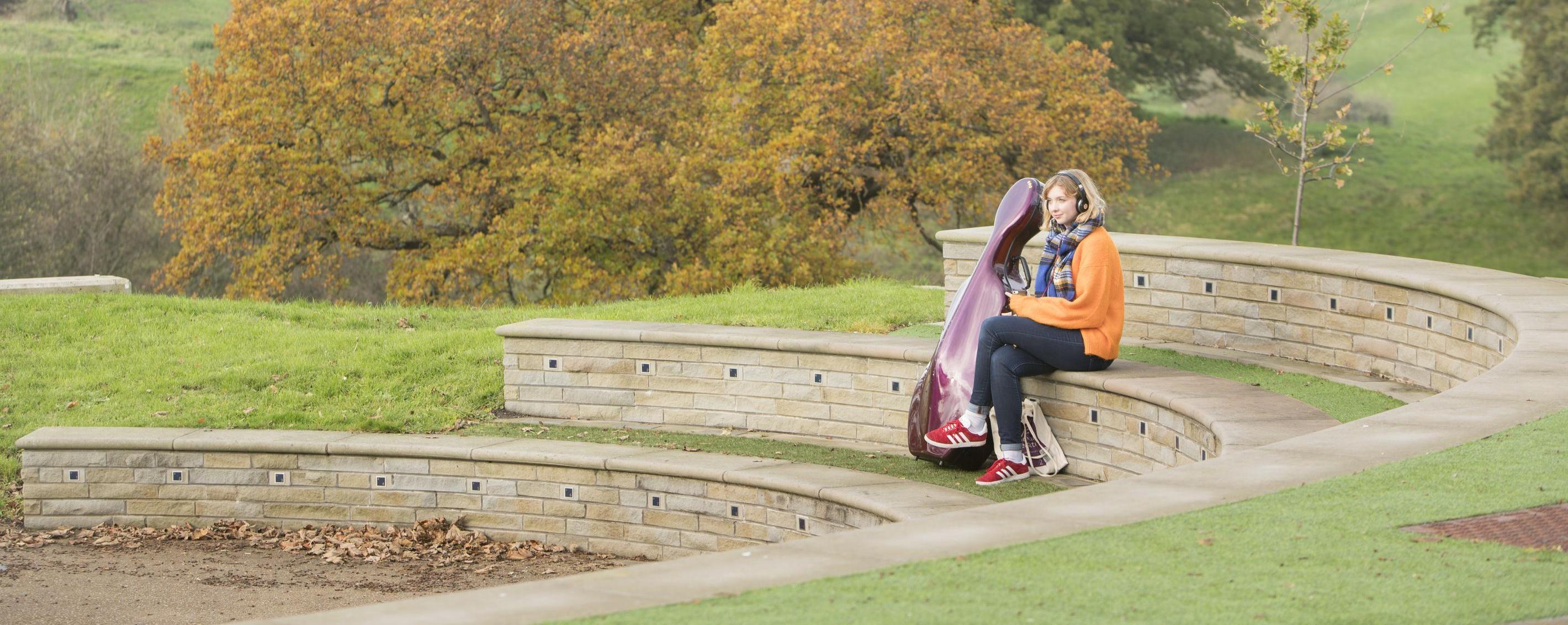 A student sits at an empty amphitheatre in a scenic location surrounded by greenery and trees