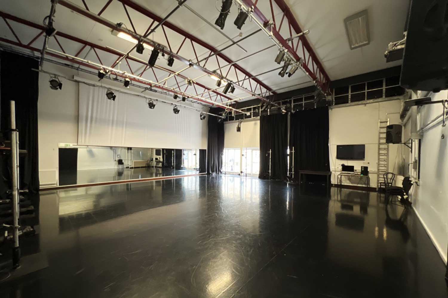 A large, empty dance studio with black flooring and curtains, lighting rig and large mirrors
