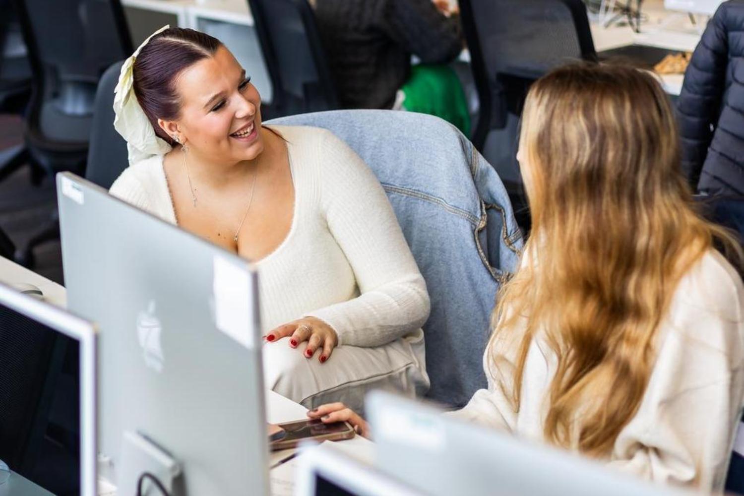 Two students turn to each other laughing and talking in a computer suite