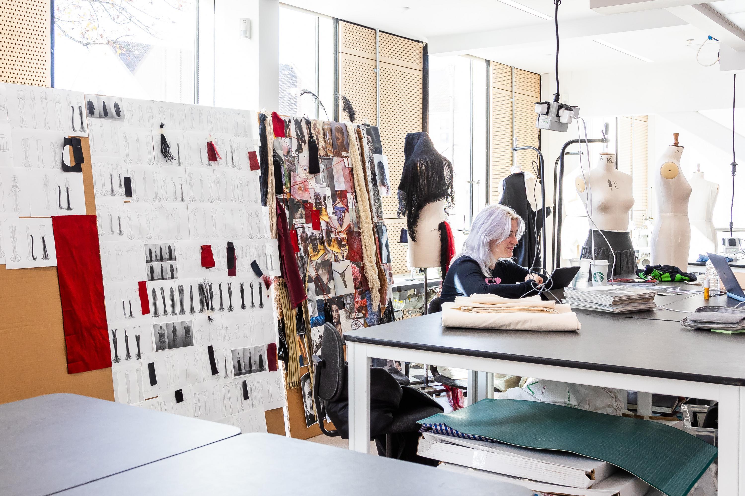 A person with lilac hair sits at a large table surrounded by sketchbooks and fabric samples on a moodboard