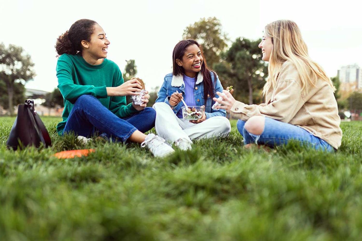 Three students eat lunch while sitting on the grass