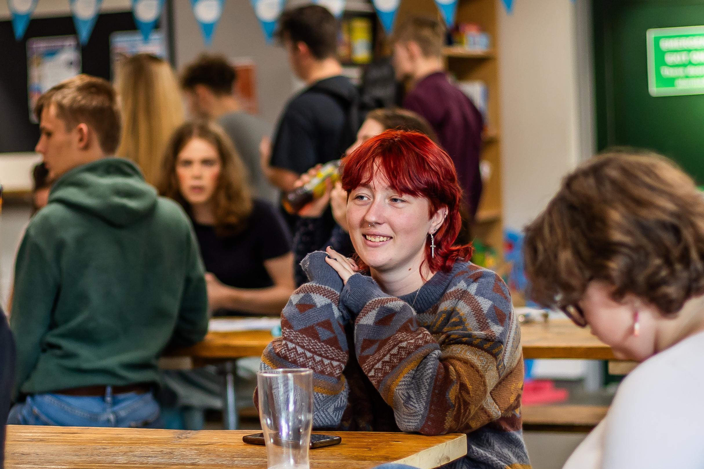 Students sit at a wooden table with people walking around behind them