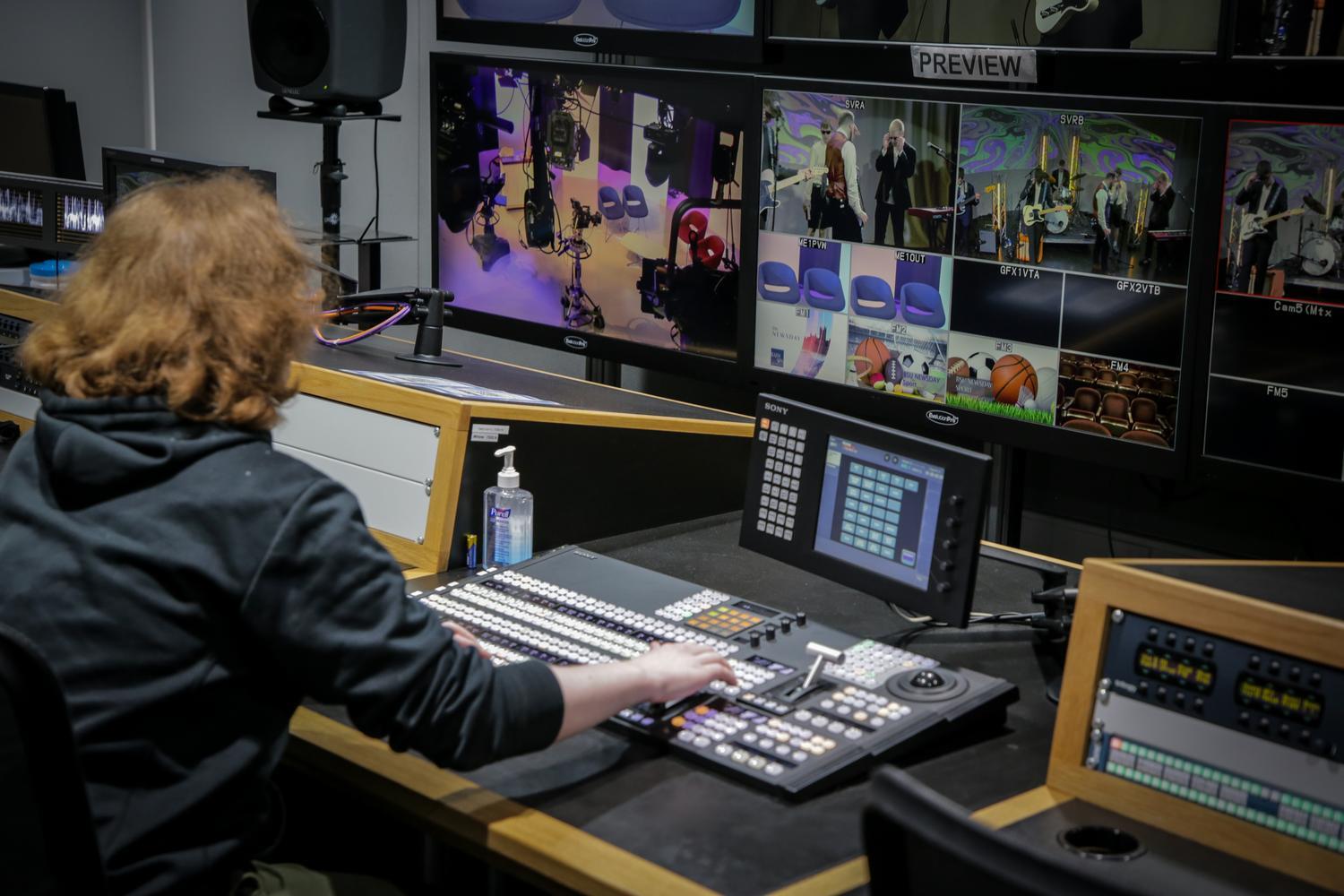 A person sits at a desk surrounded by large screens showing different angles of the TV studio