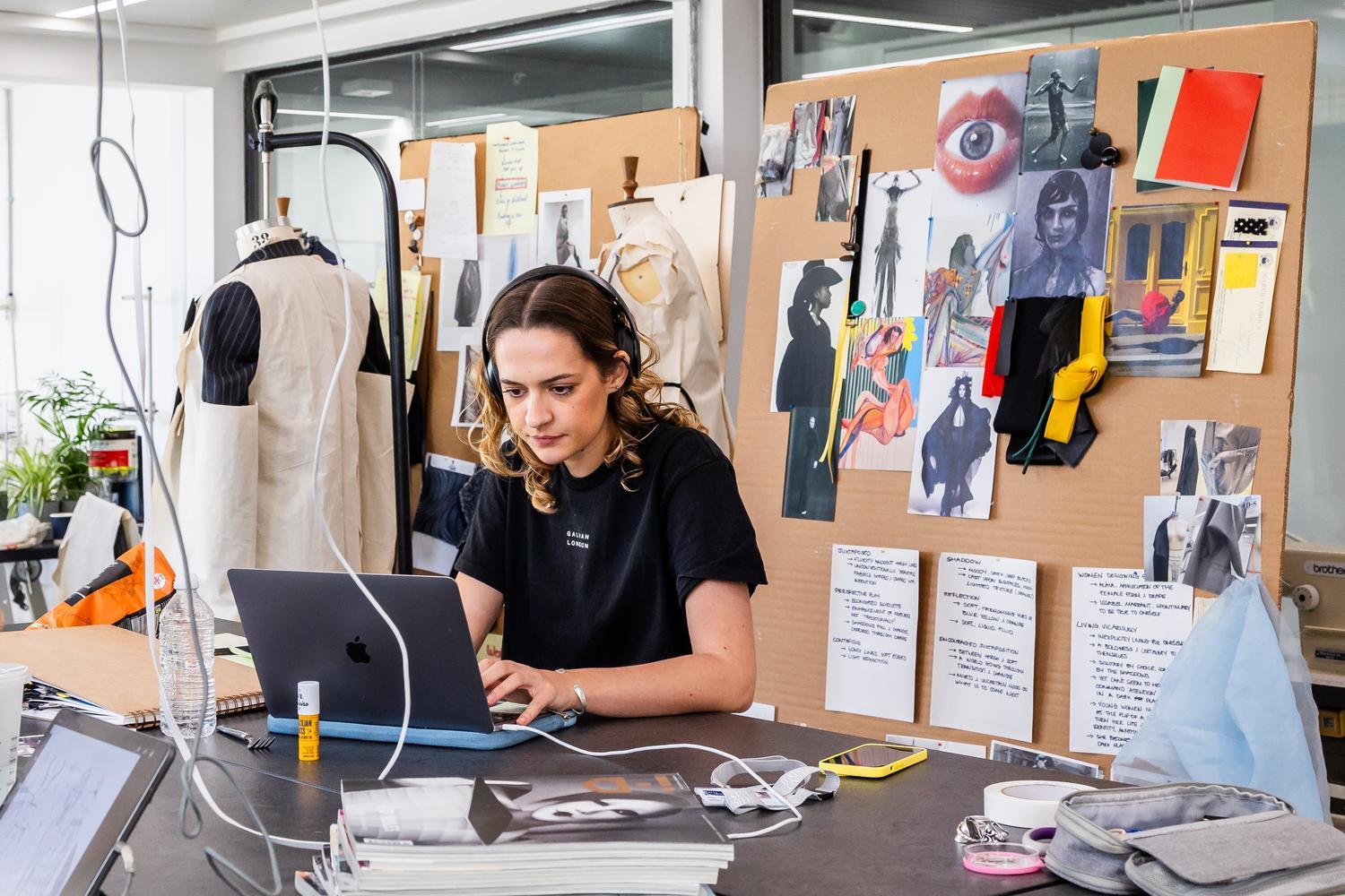 A student types on a laptop while surrounded by fashion designs