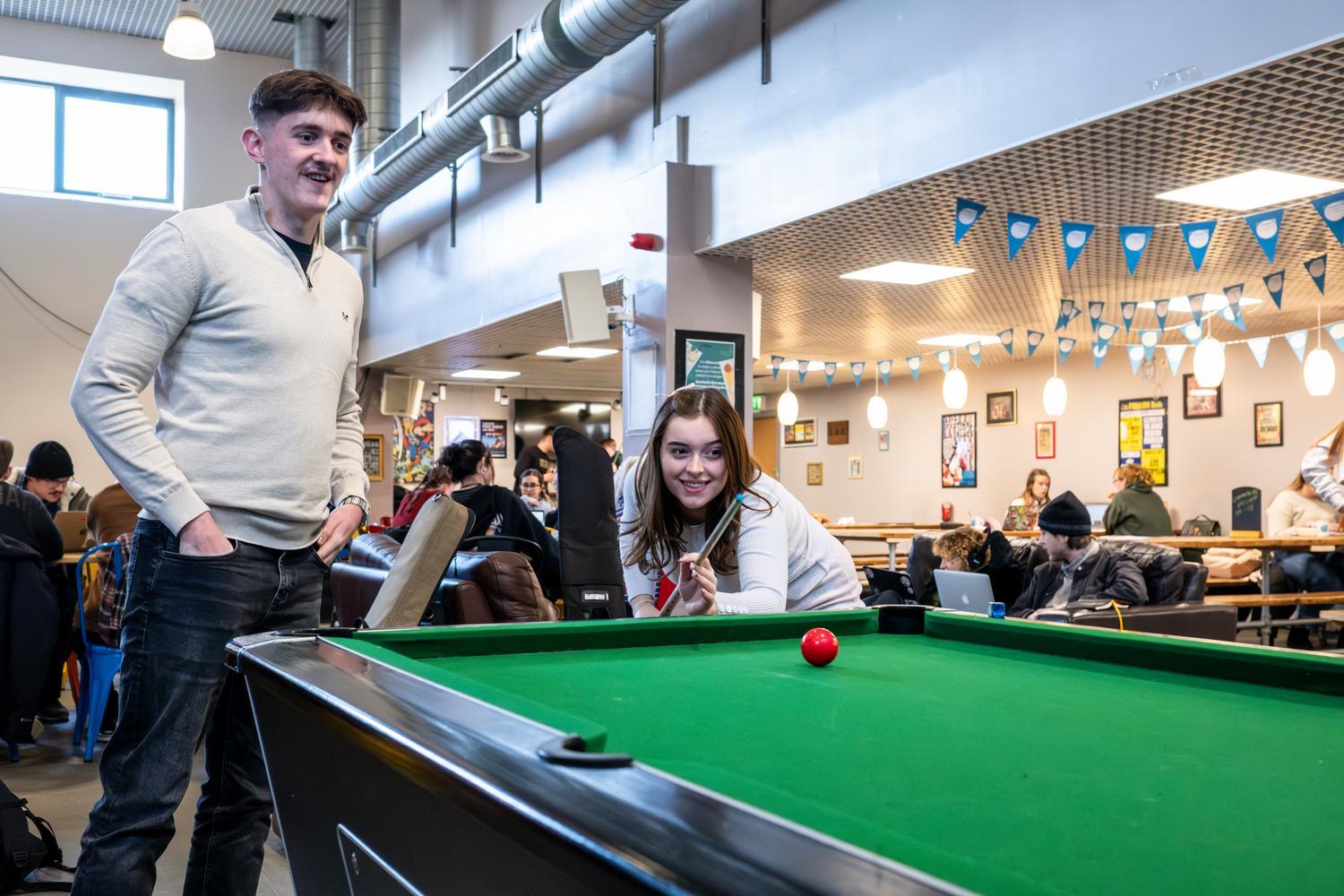 Two students play pool inside a social common room area