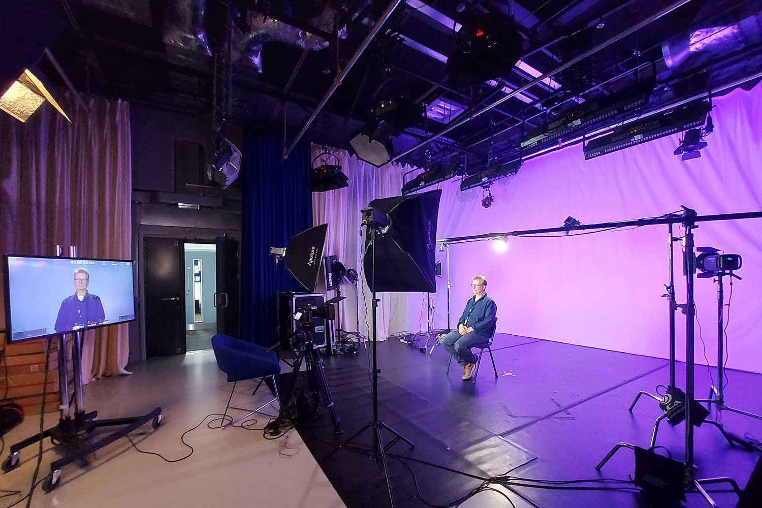 A TV studio with purple lighting and a person sat on a chair in front of the camera