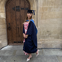 A Bath Spa student wearing graduation gown stands in front of a yellow brick wall.
