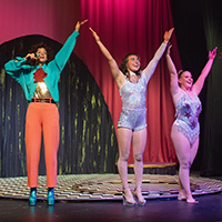 Three performers stand on a stage with arms outstretched.