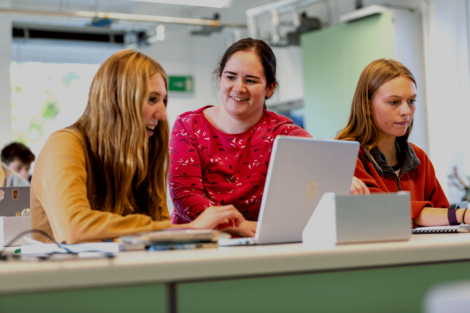 Two students sit at desk on their laptops. In the middle of the two students a lecturer stands talking to the student on the left.