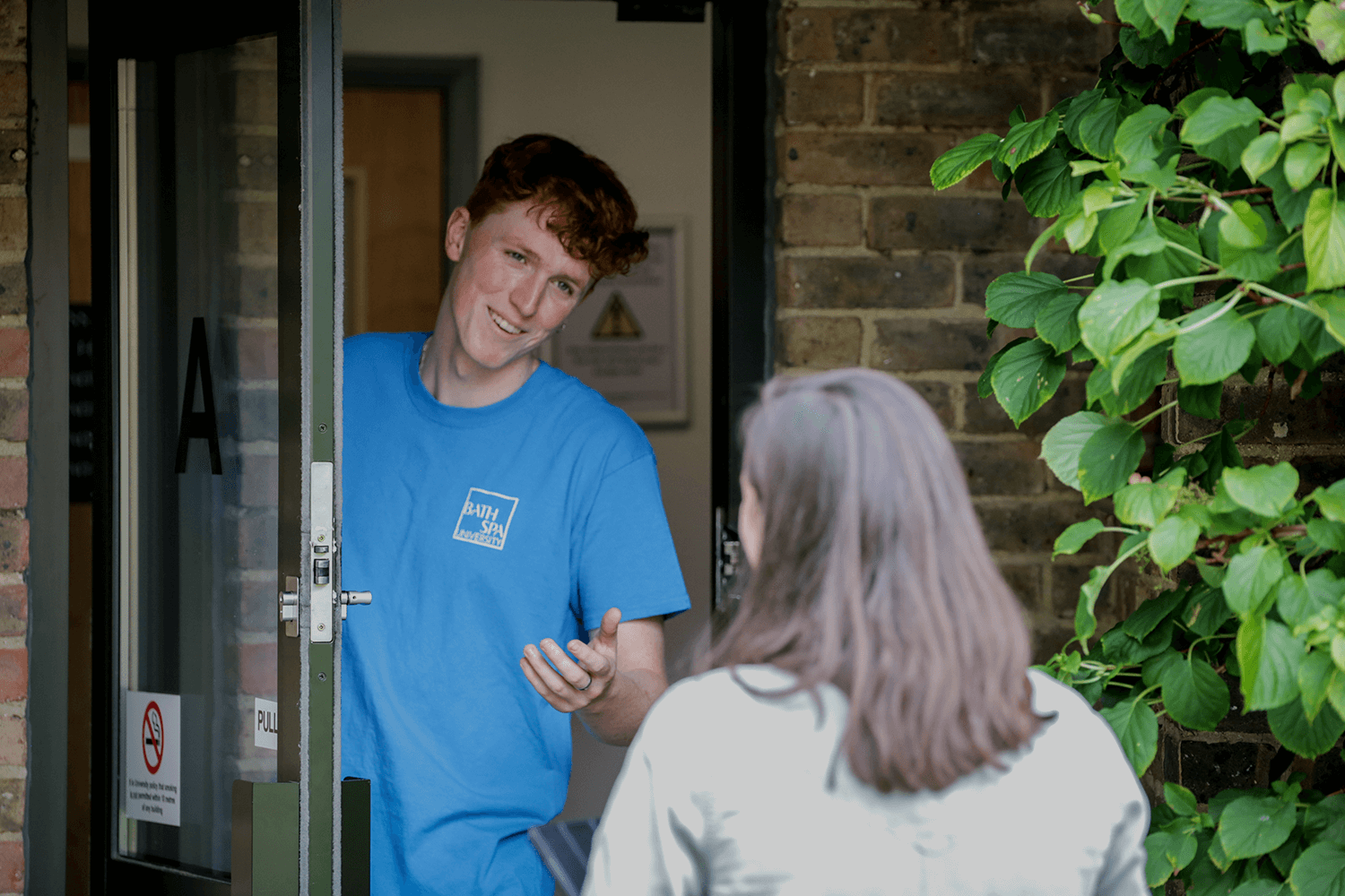 A white male student with red hair and wearing a blue shirt stands in the doorway of a hall of residence. He is smiling and talking to another student,