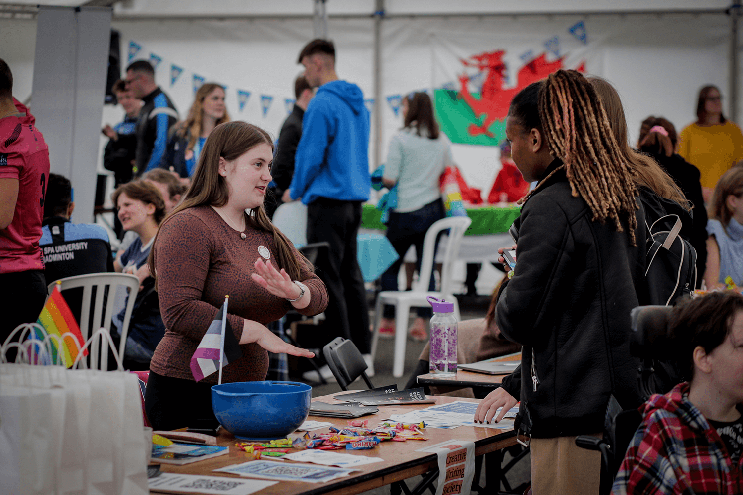 a member of the creative writing society at bath spa university stands behind their table at the activity fair. They are talking to a interested student who is stood the other side of the table