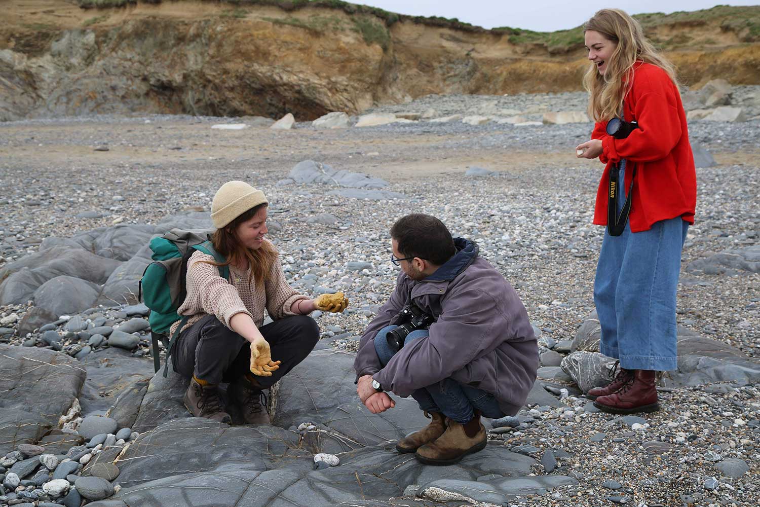 Three people crouch on the sand to examine various materials