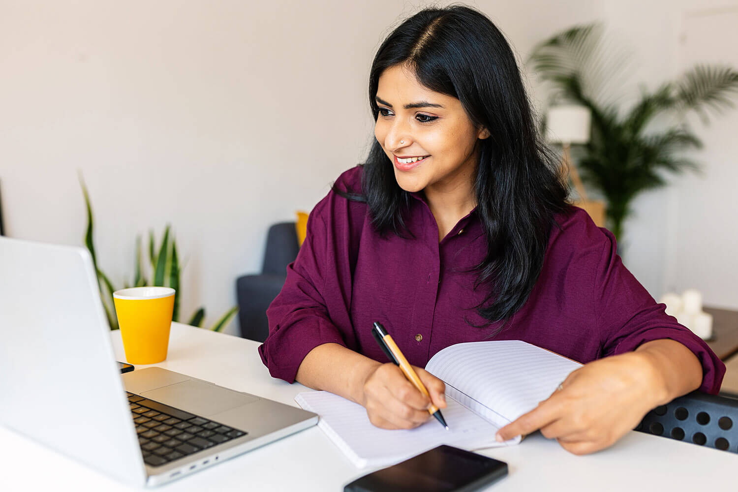 Smiling student taking notes while using laptop at home