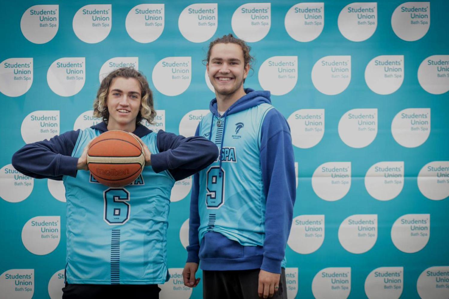 Two students stand in front of the SU backdrop, holding a sports ball