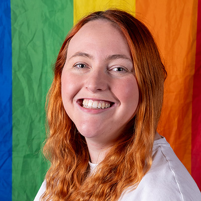 A smiling Bath Spa University staff member sitting in front of a colourful flag.