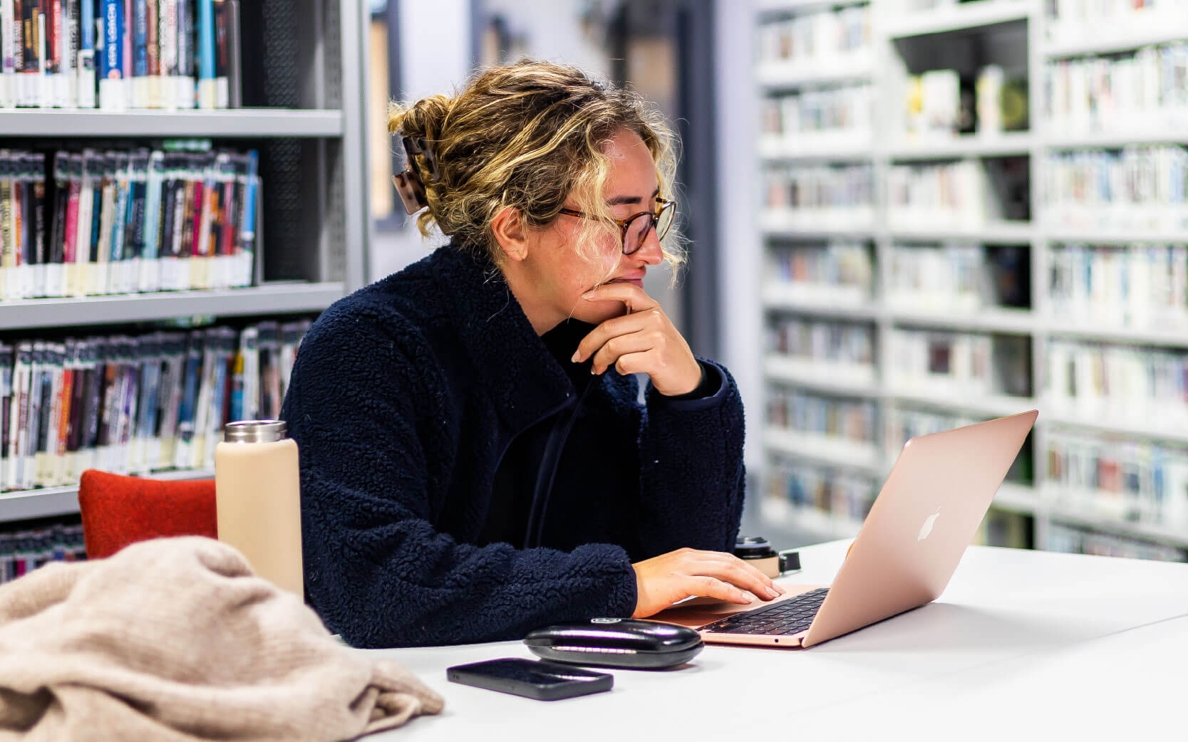 Student working on a rose gold laptop in a library, with shelves of books behind her and a phone, water bottle and cardigan nearby on the table